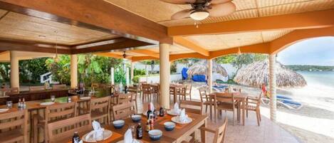 Un restaurant situé en bord de mer, avec des tables et des chaises en bois, un ventilateur de plafond et une vue sur la plage et l’océan.