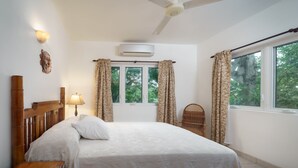 Een slaapkamer met een bed, nachtkastje, lamp, airconditioning, raam met gordijnen en uitzicht op groen buiten.