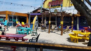 An outdoor seating area with tables and chairs, a beachside restaurant with a yellow facade, and a wooden structure with hammocks. - Romantic Beach Getaway For 2 In Ocean City MD (Ocean City)