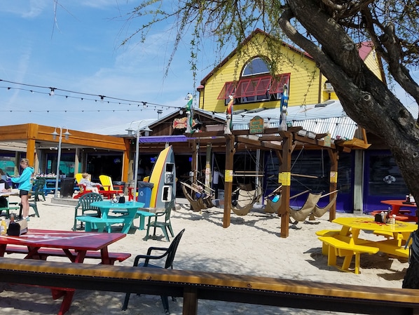 An outdoor seating area with tables and chairs, a beachside restaurant with a yellow facade, and a wooden structure with hammocks.