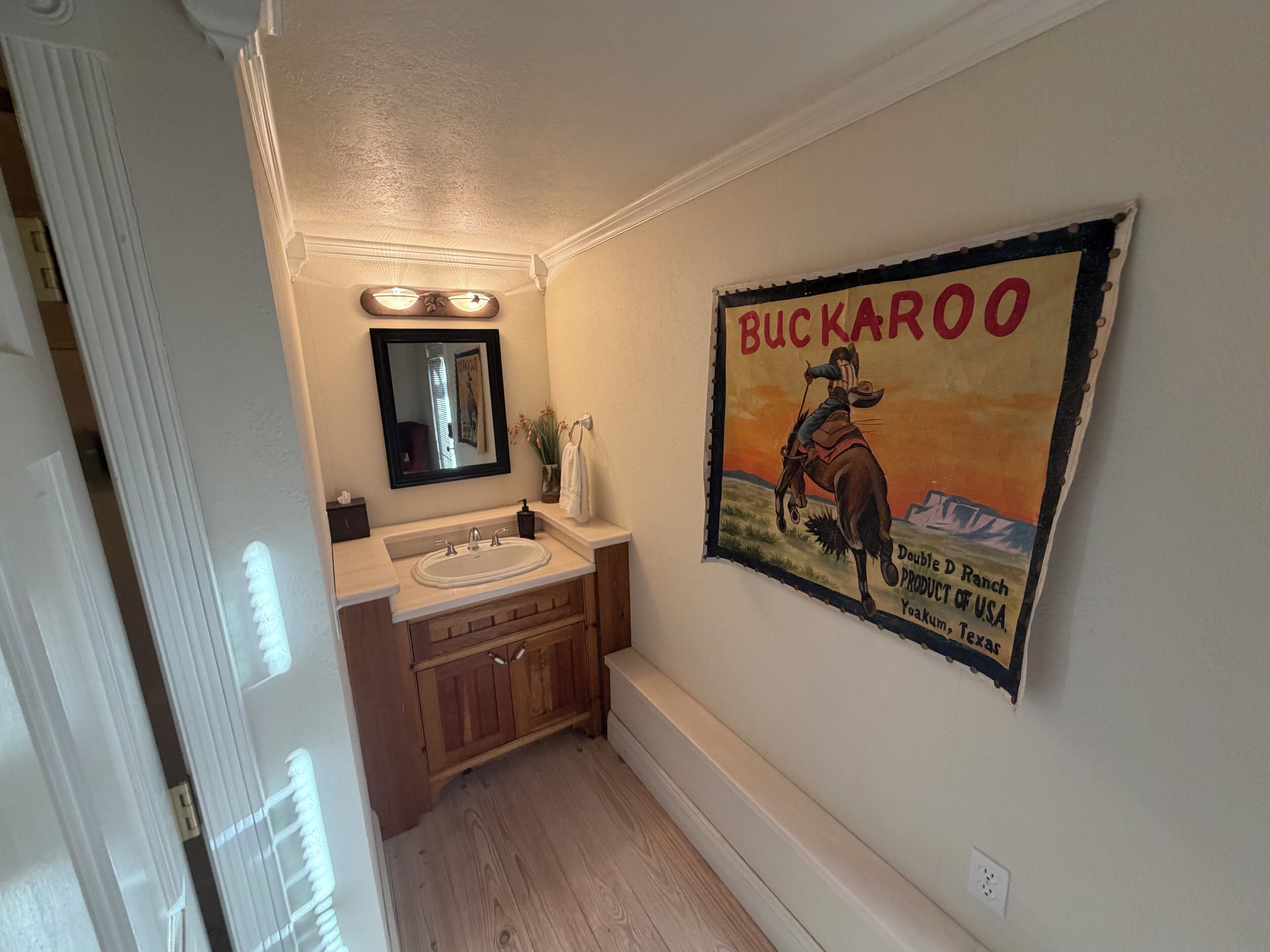 Un baño con tocador de madera, un lavamanos y un espejo. Hay una lámpara de pared sobre el espejo y un cartel con un vaquero montando a caballo.