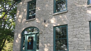 Ein zweistöckiges Steinhaus mit grüner Tür und Fenstern, umgeben von Bäumen und einer Rasenfläche.