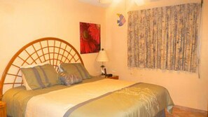 Un dormitorio con cabecera de madera, cama con colcha verde, dos almohadas, mesita de noche con lámpara y un tapiz en la pared.