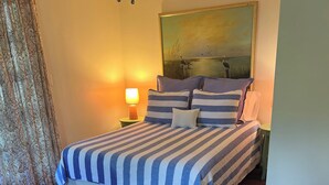 Un dormitorio con una cama grande, una mesita de noche con lámpara, un cuadro en la pared y una ventana con cortinas.
