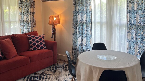 Una habitación con un sofá rojo, una mesa redonda con sillas, cortinas estampadas y un ventilador de techo.