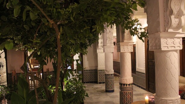 A traditional interior with ornate columns, a wooden table with a candle, and lush greenery.
