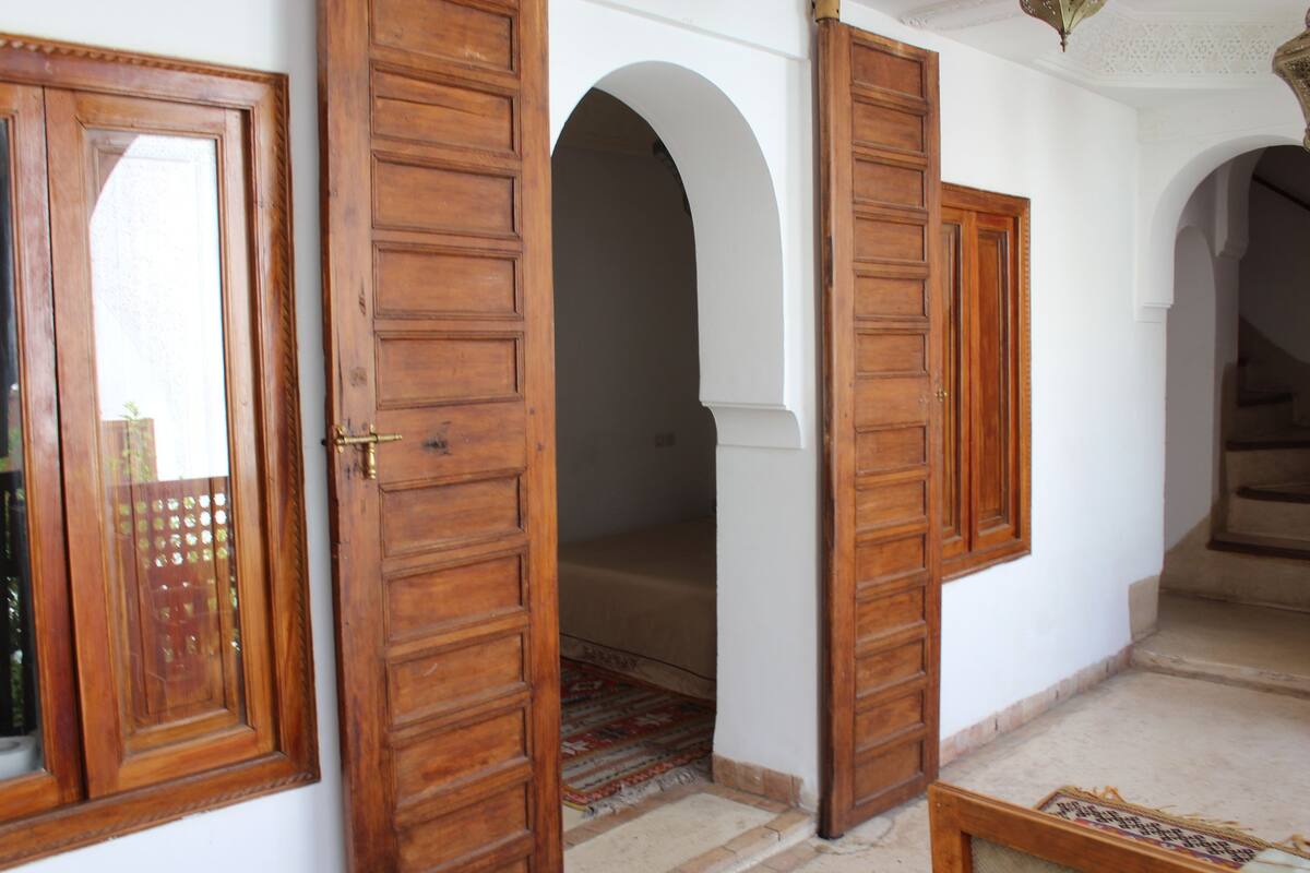 Un couloir avec des portes et des arches en bois, qui mène à une pièce contenant un lit et un tapis à motifs.