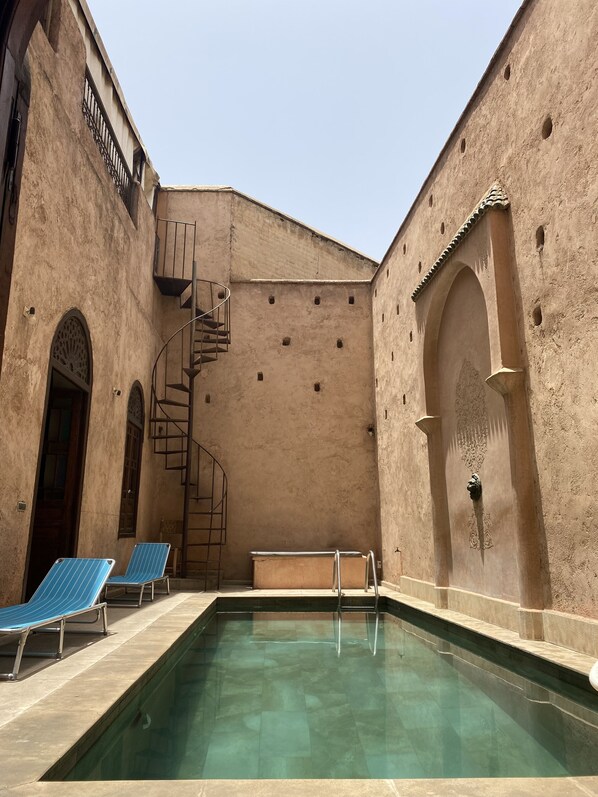 Una piscina con escalera espiral, sillas reclinables azules y un muro con detalles decorativos.