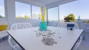 A modern dining area with a white table, clear glass vase with yellow straws, scattered stones, and white chairs. - Villa Tramonto (Seget)