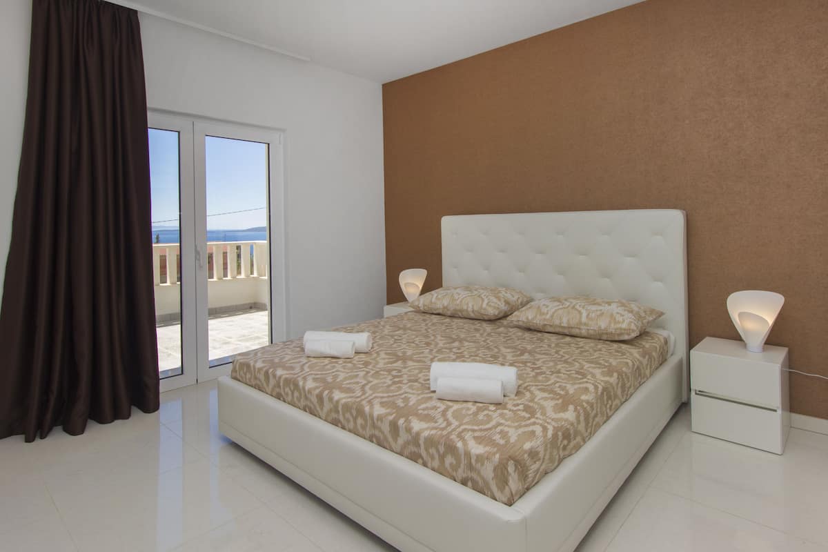 A modern bedroom with a large bed, a nightstand, and a view through a sliding door to the outside.