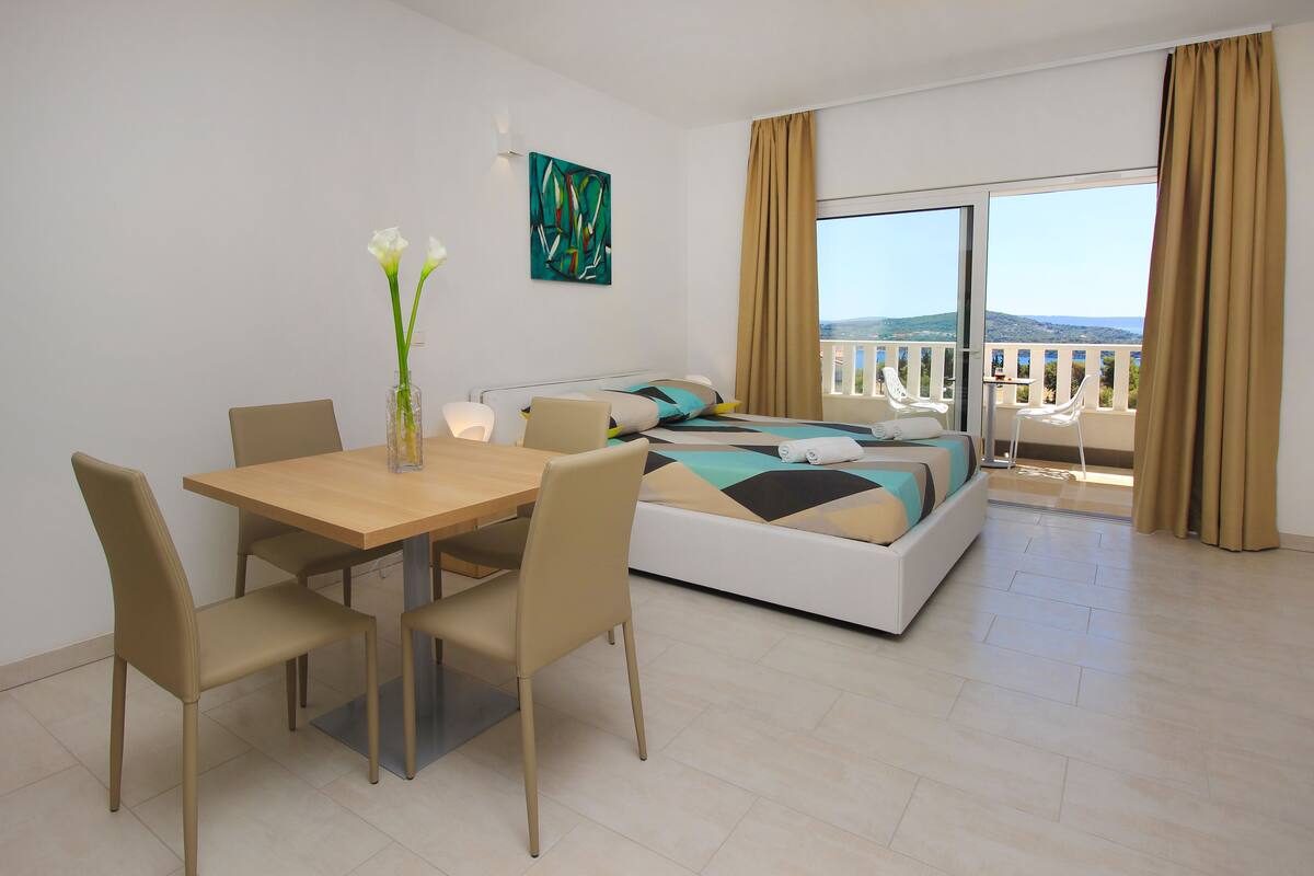A modern hotel room with a bed, a dining table with chairs, a vase with flowers, and a balcony with a view.