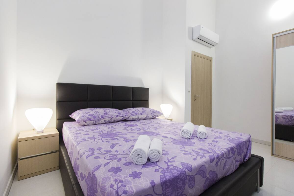 A neatly made bed with a floral bedspread, two bedside lamps, and a wall-mounted air conditioner.