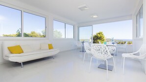 A modern, bright room with a white sofa, white chairs, and large windows offering a view of greenery and a distant landscape. - Villa Tramonto (Seget)