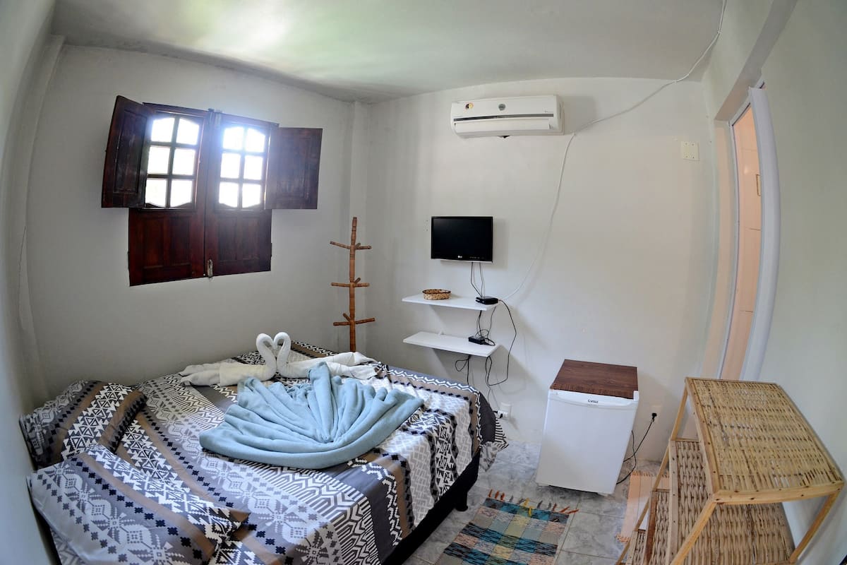 A bedroom with a bed, a TV, an air conditioner, and a small white refrigerator.