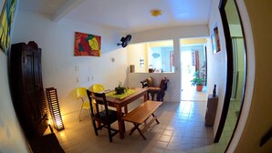A dining area with a wooden table, chairs, and a bench. There is a colorful painting on the wall, a ceiling fan, and a potted plant in the background. - Residencial Porto de Galinhas (Ipojuca)