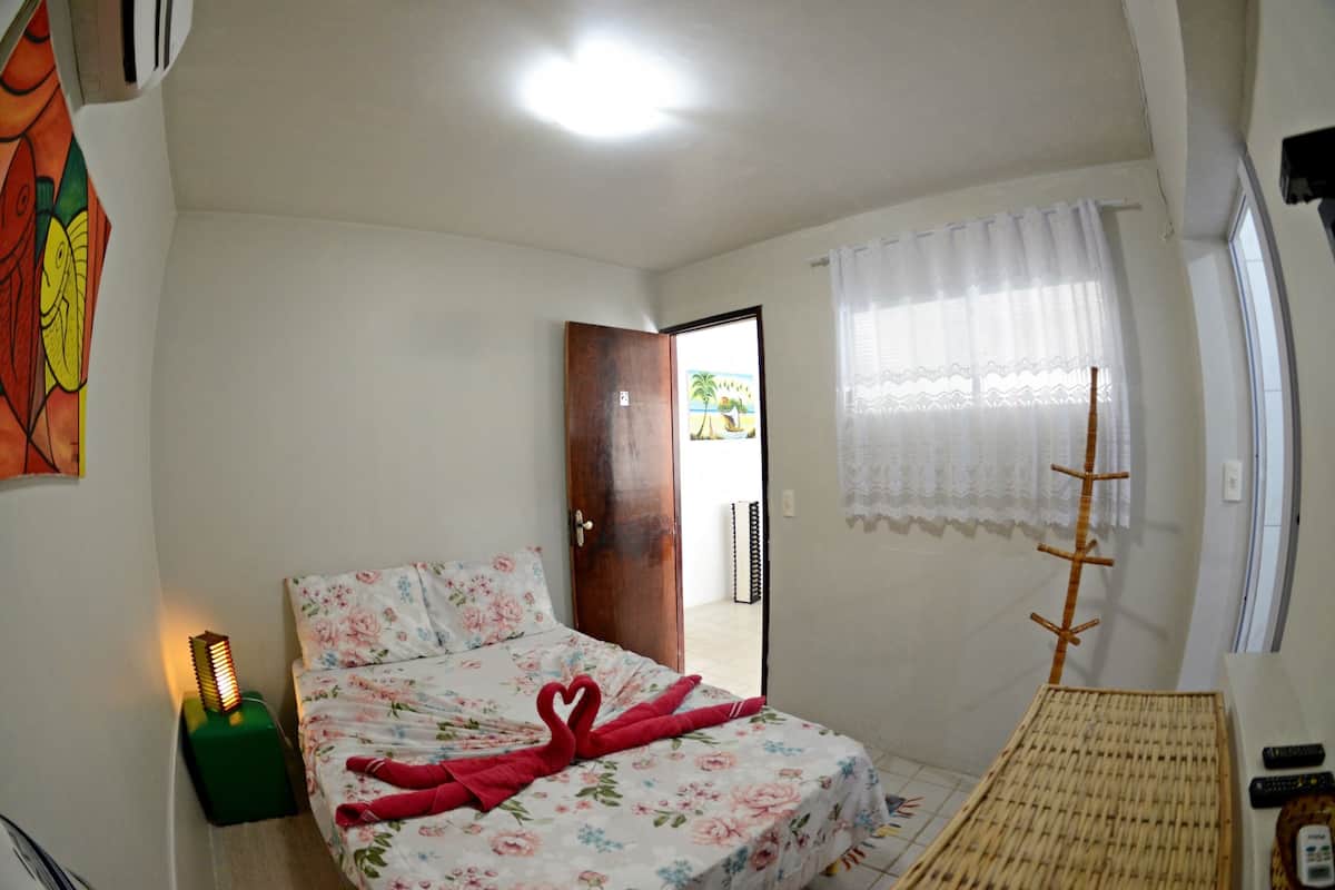 A bedroom with a bed, a nightstand, a lamp, a window with a curtain, and a door leading to another room.