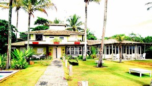 Un edificio de dos plantas con tejado de tejas, piscina y jardín con palmeras y sillas.
