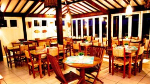Un restaurante con varias mesas y sillas de madera, amplios ventanales y suelo de baldosas.