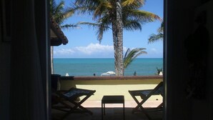 Un acceso abierto a un balcón con dos sillones de relax y una pequeña mesa, con vistas a una playa con palmeras y el océano.