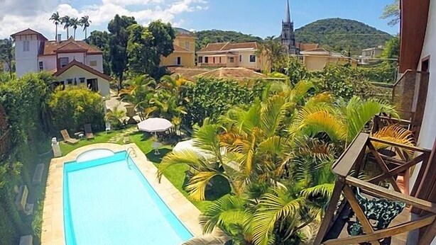 A view of a swimming pool surrounded by lush greenery and palm trees, with buildings and a church in the background. - Pousada Imperial Koeler (Petrópolis)
