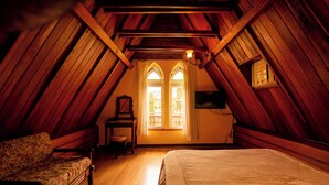 A cozy attic bedroom with a wooden sloped ceiling, a bed, a chair, and a desk. - Pousada Imperial Koeler (Petrópolis)