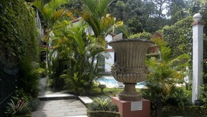 A garden pathway leading to a large decorative urn, surrounded by lush greenery and a white building in the background. - Pousada Imperial Koeler (Petrópolis)