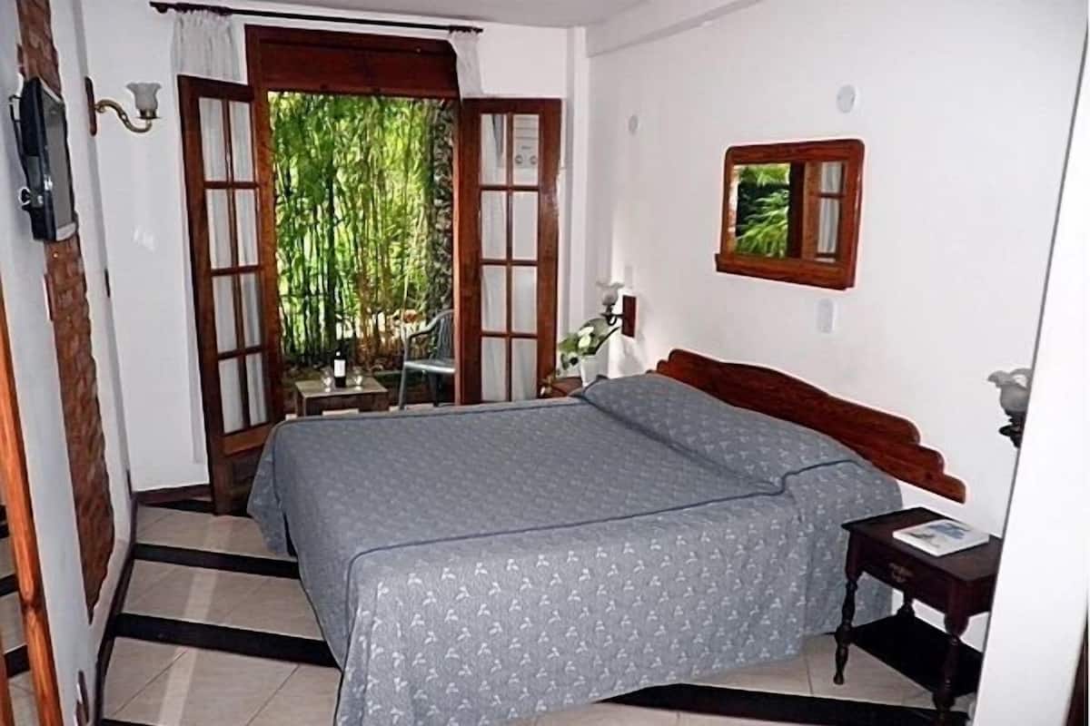 A bedroom with a bed, a nightstand, a window with a view of greenery, and a door leading outside.