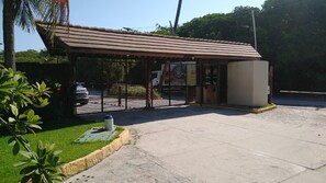 A covered outdoor area with a car parked inside, a small building with a metal roof, and a concrete floor. - Nannai Residence Apartments - Muro Alto (Porto de Galinhas - PE) (Ipojuca)
