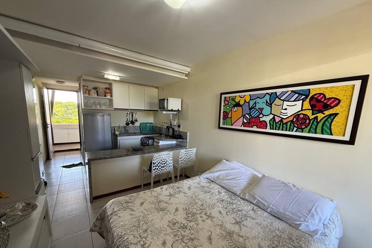A bedroom with a bed, a painting, and a kitchenette.