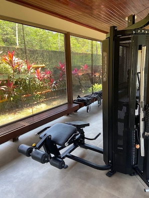 A gym with exercise equipment, benches, and a view of a garden through large windows.