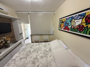 Ein Schlafzimmer mit einem Bett, einem Fernseher, einem Schreibtisch mit einer Blumenvase und einem farbenfrohen, abstrakten Gemälde an der Wand.