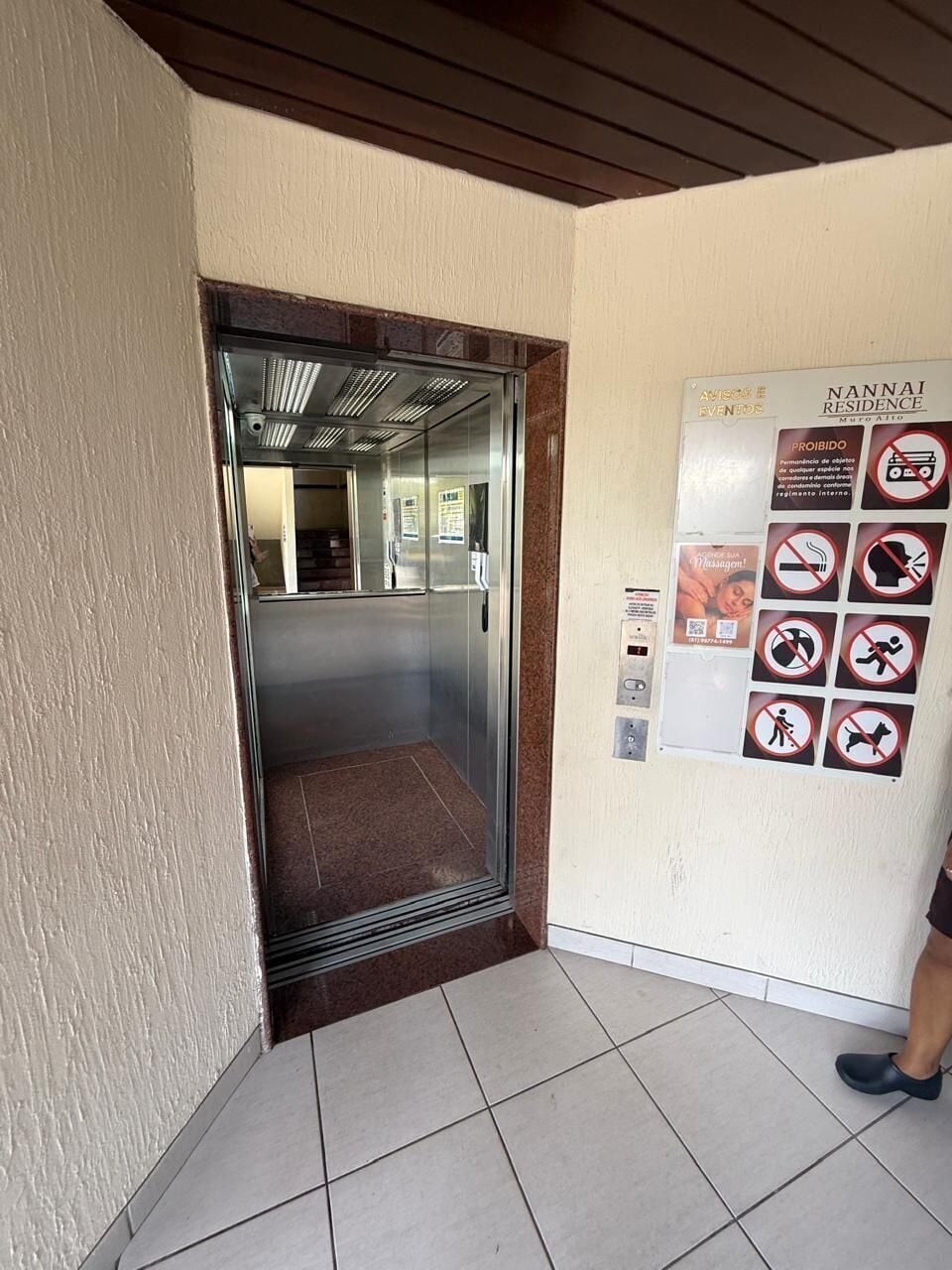 Um elevador com porta de vidro, uma placa com itens proibidos e chão de azulejos.