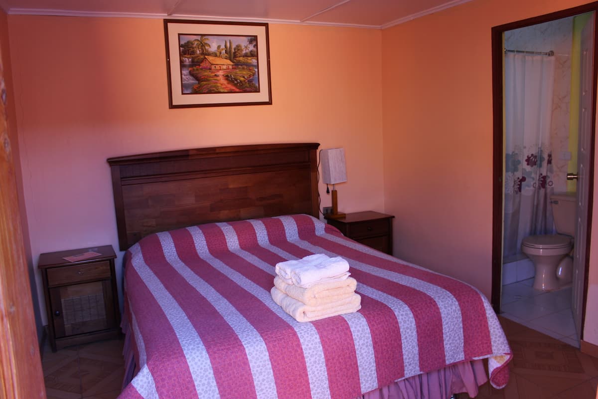 A neatly made bed with a striped bedspread and folded towels, a wooden headboard, a nightstand with a lamp, and a painting on the wall.