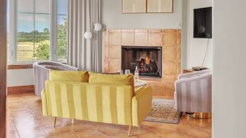 Un salon avec un canapé jaune, une cheminée et une grande fenêtre donnant sur un paysage verdoyant.