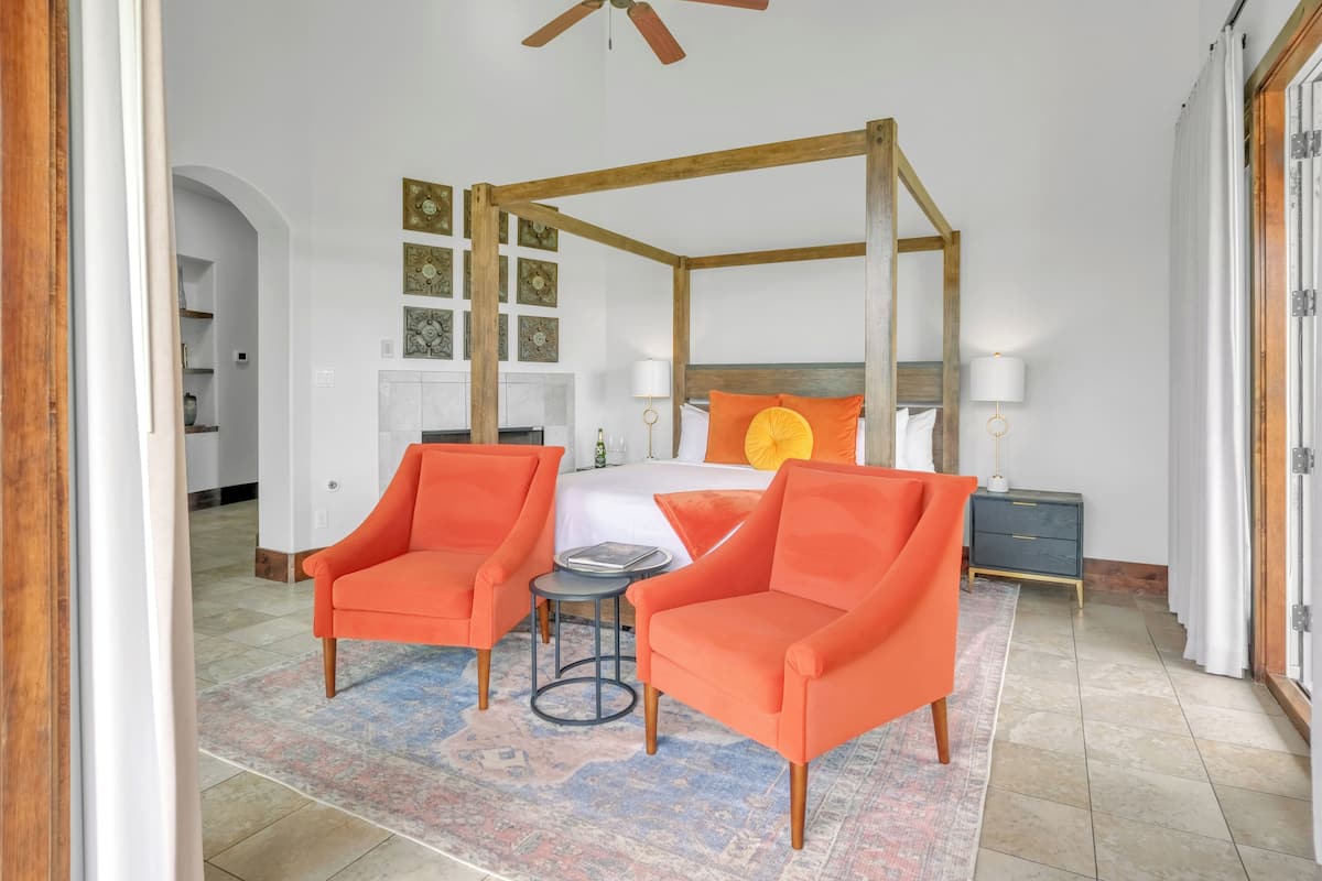 A bedroom with a four-poster bed, two orange armchairs, a small round table, and a nightstand with a lamp.