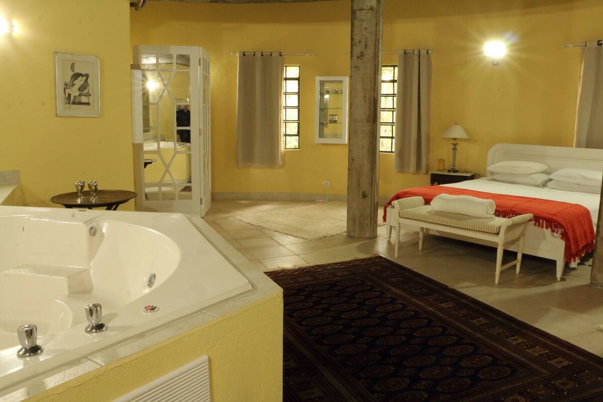 Une chambre d’hôtel avec une grande baignoire, un lit recouvert d’une couverture rouge, un banc et un tapis à motifs.