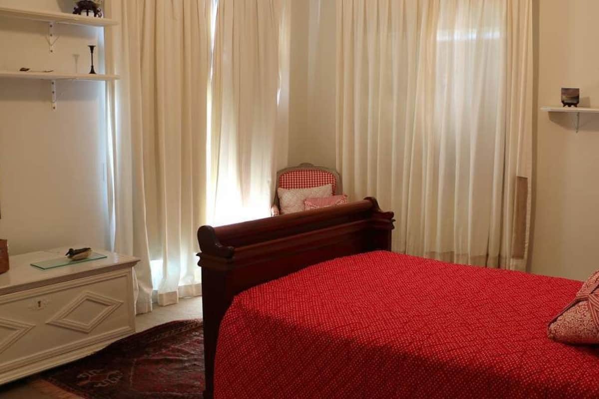 Une chambre avec un lit en bois, une couvre-lit rouge et une chaise à carreaux rouges et blancs. On y trouve des rideaux blancs, une commode blanche et une étagère avec des objets décoratifs.