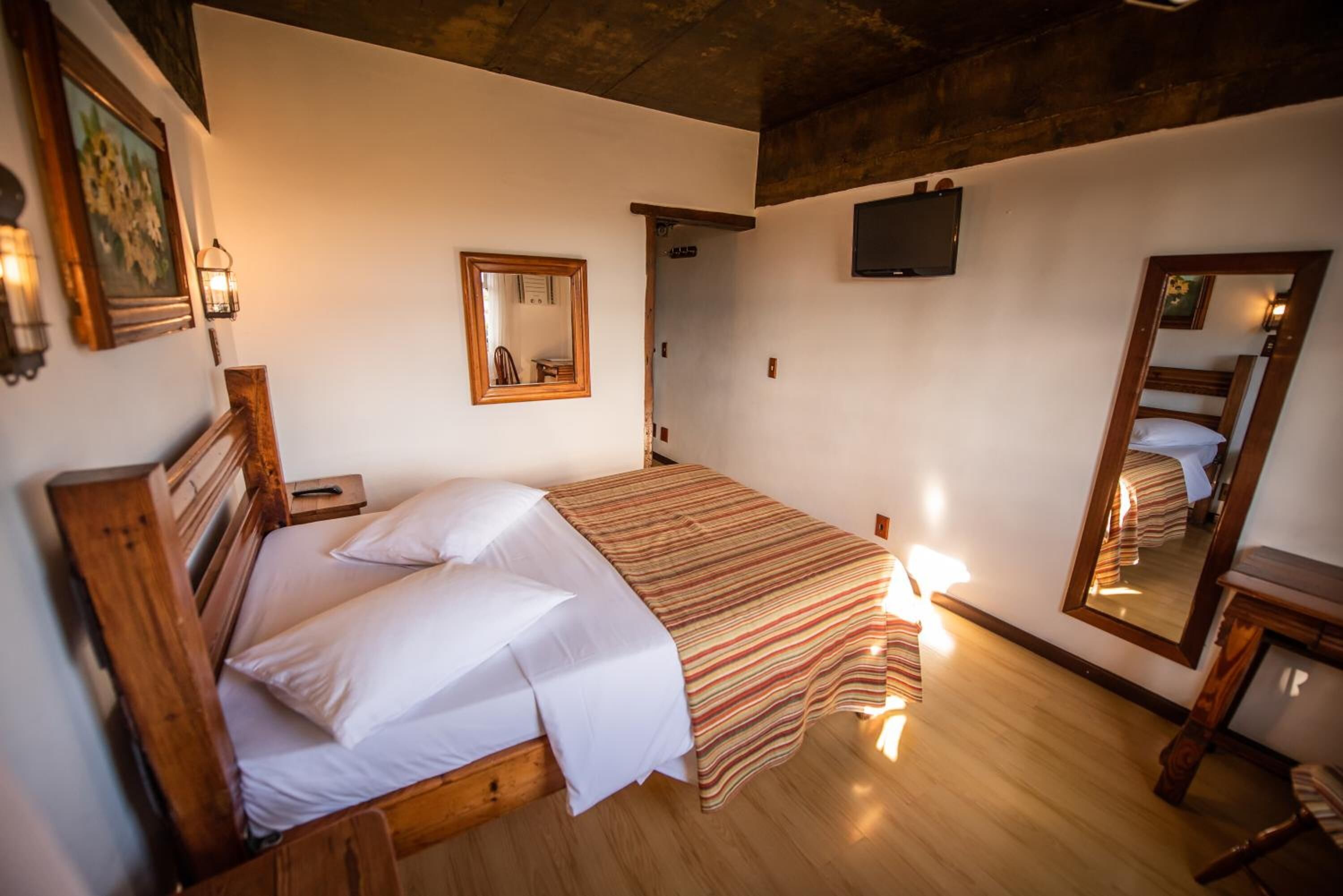 Quarto com cama de madeira, espelho, televisão e porta que dá para outro cômodo.