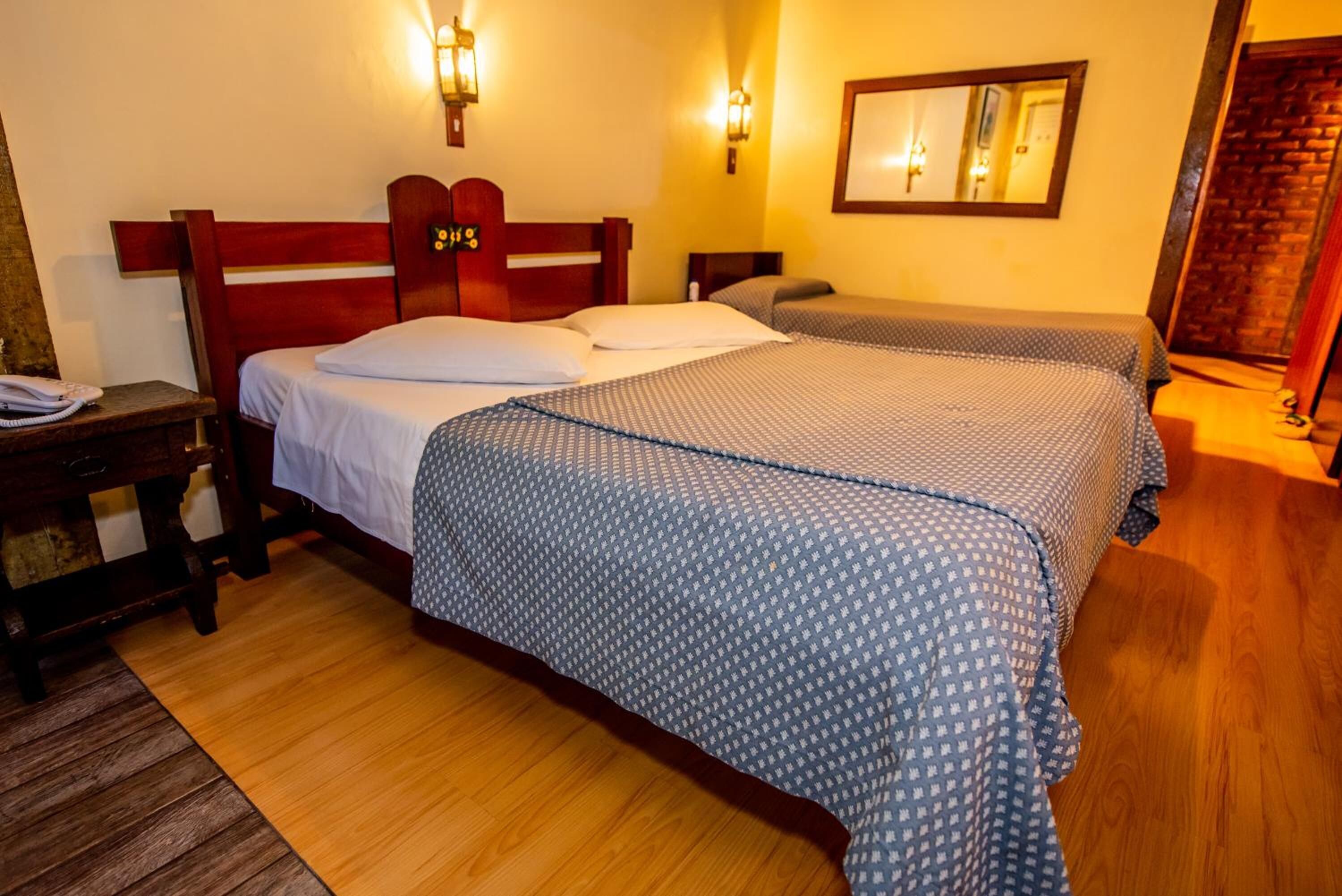 Quarto de hotel com duas camas, cabeceiras de madeira, piso de madeira, mesa de cabeceira com telefone e luminárias fixadas na parede.
