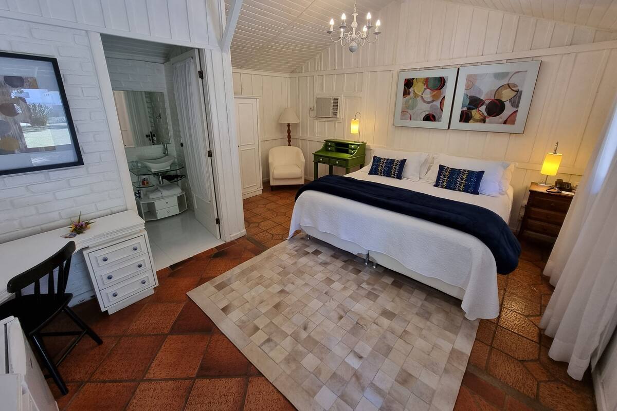 A bedroom with a bed, bedside tables, a desk, a chair, a mirror, and a bathroom visible through an open door.