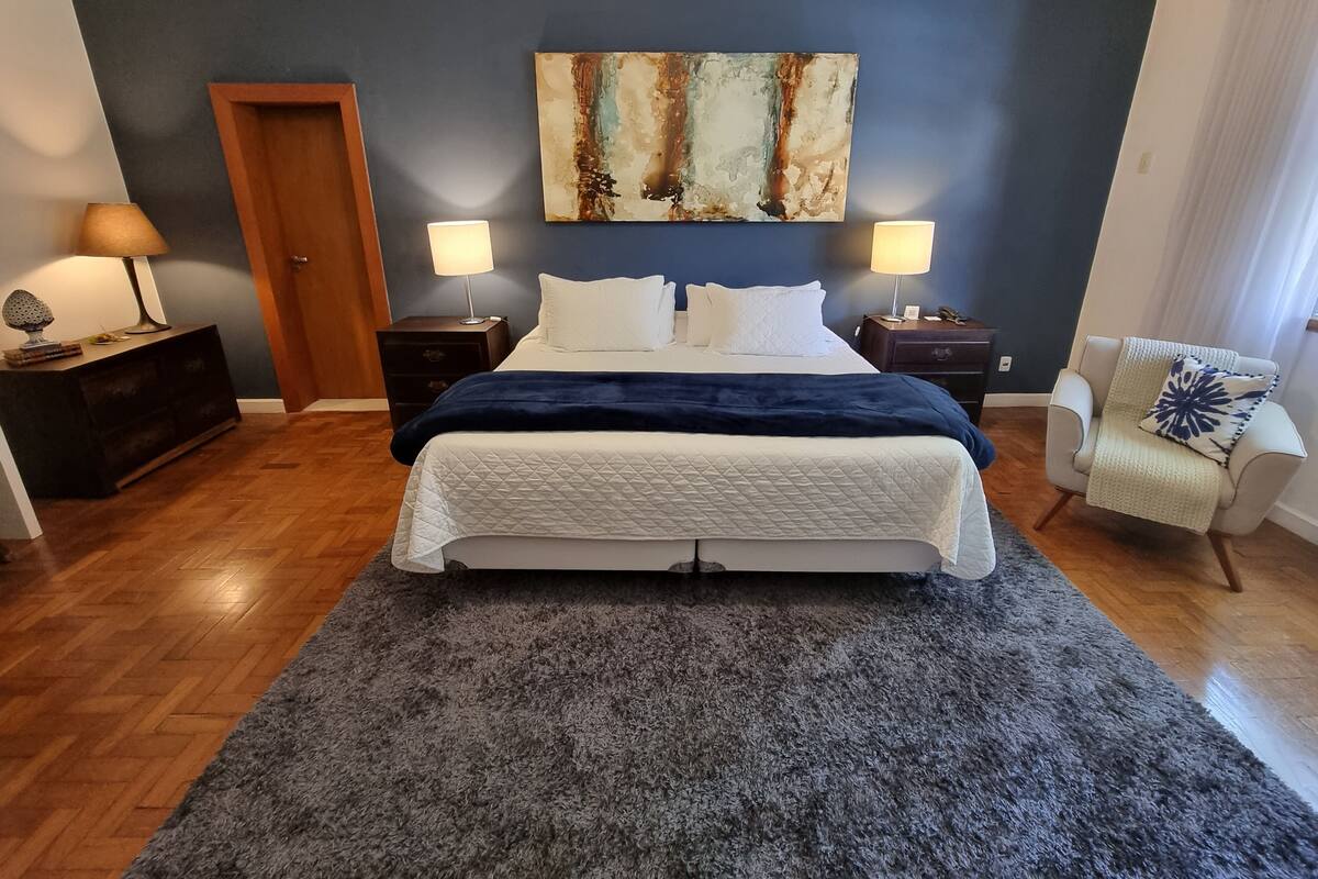 A bedroom with a large bed, two bedside tables with lamps, a chair, a wooden door, and a painting on the wall.