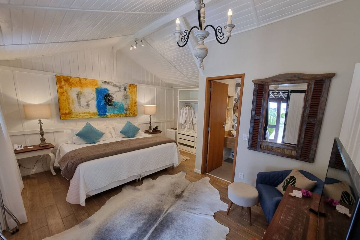 A spacious bedroom with a large bed, a desk, a chair, a sofa, a mirror, and a painting on the wall.