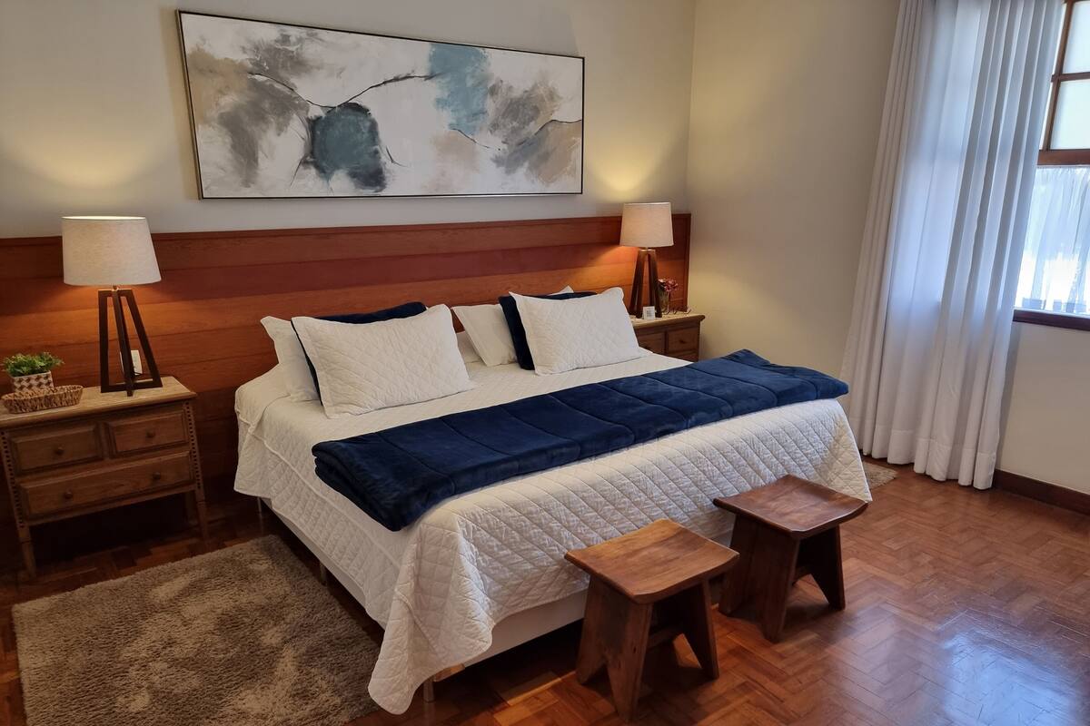 A neatly arranged bedroom with a large bed, bedside tables, a nightstand, a small wooden stool, a rug, and a painting on the wall.