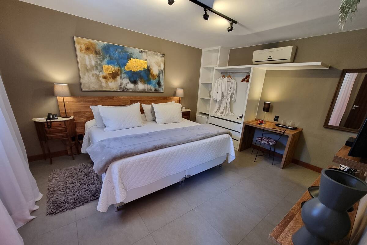 A modern hotel room with a large bed, a desk, a wardrobe, and a painting on the wall.
