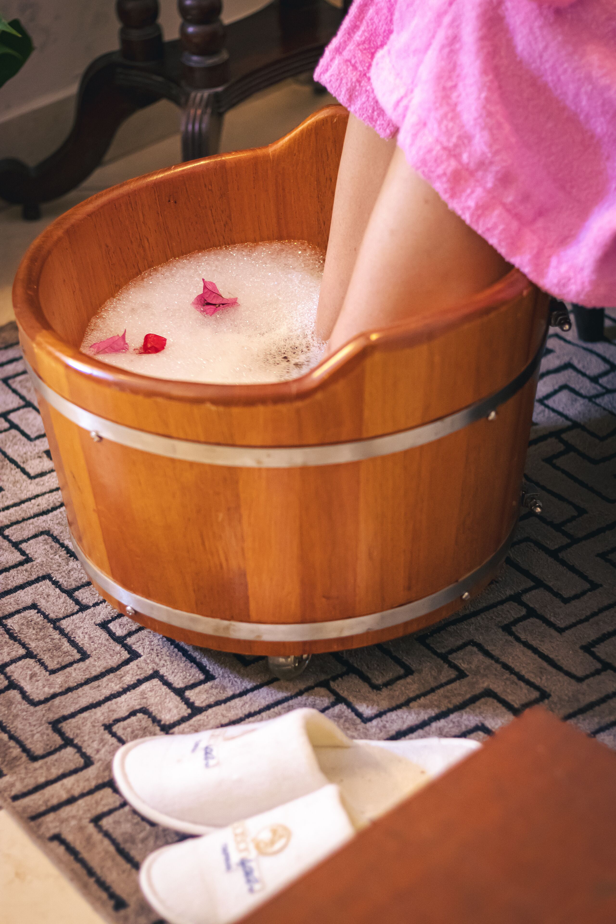 A wooden tub filled with bubbles and rose petals, placed on a patterned rug.