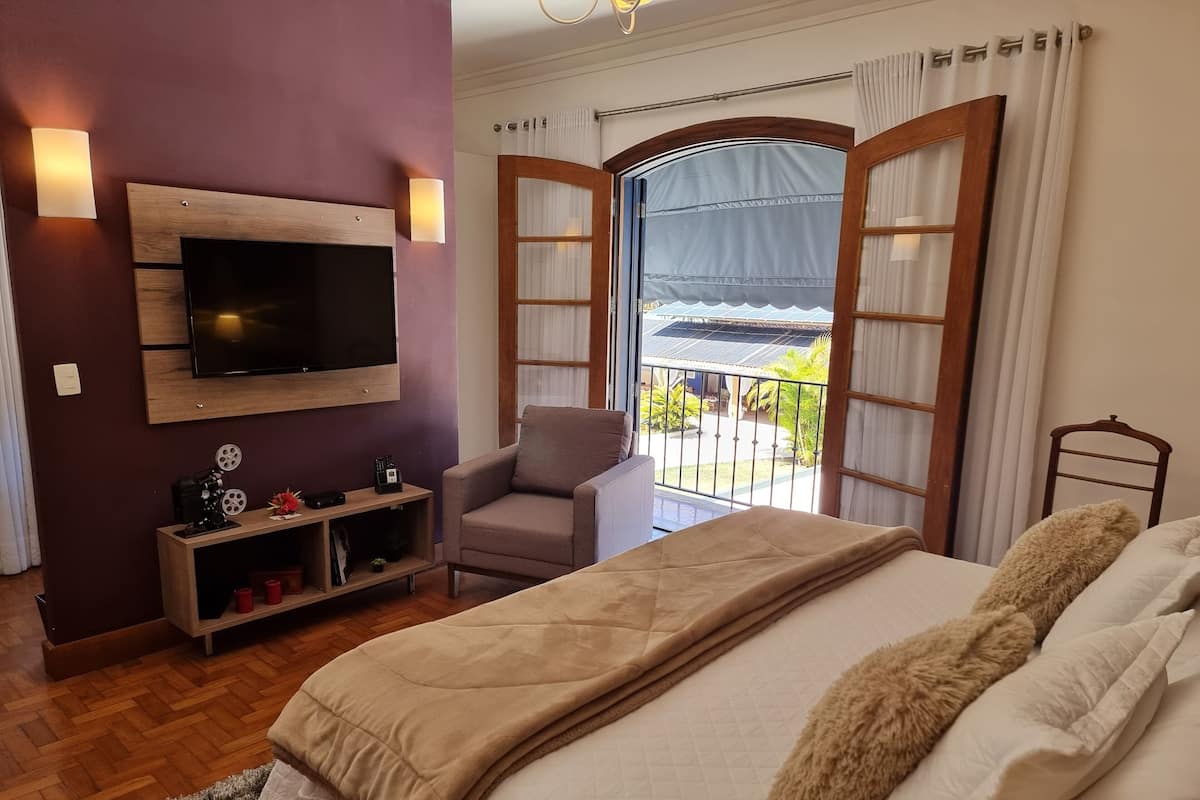 A bedroom with a bed, a TV mounted on the wall, a chair, a small shelf with decorative items, and a balcony with curtains.