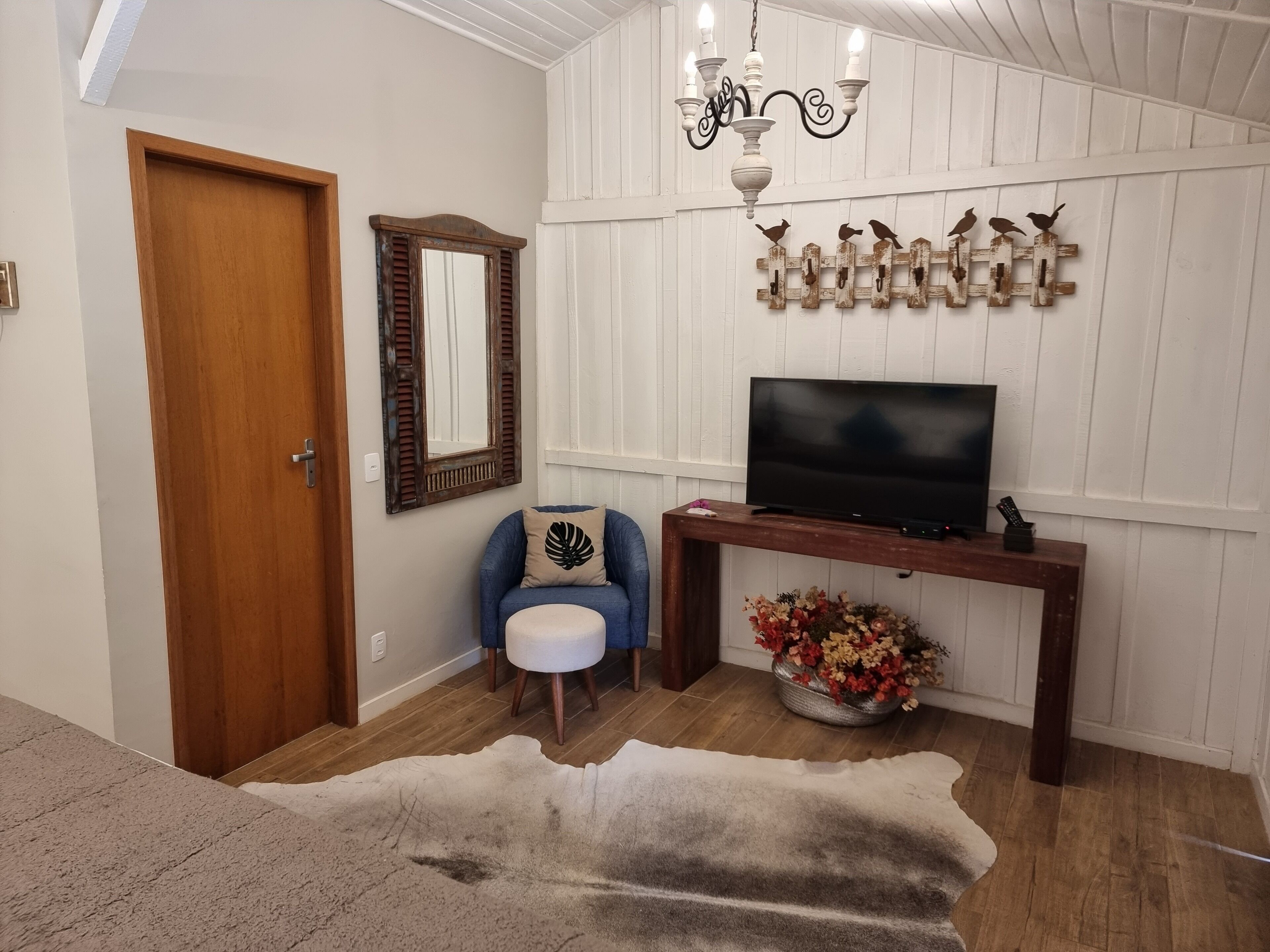 Uma sala de estar com porta de madeira, espelho, poltrona azul, televisão sobre um console de madeira e parede decorativa com elementos de madeira.