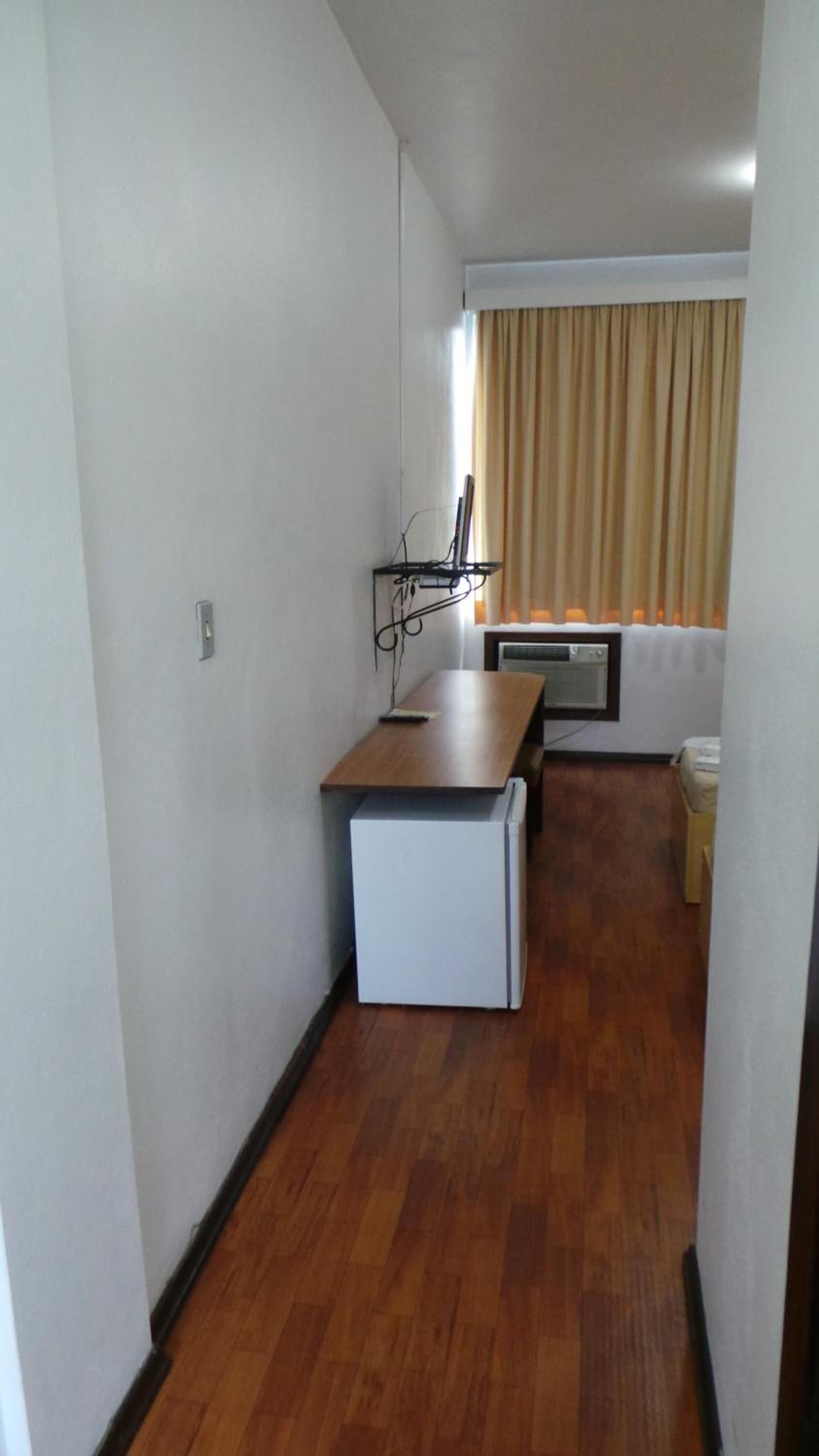 A narrow hallway with a wooden floor, a white refrigerator, a small table with a chair, a wall-mounted air conditioner, and a window with beige curtains.