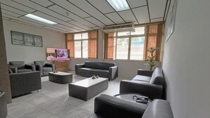 A modern lounge area with multiple sofas, a flat-screen TV, and large windows with curtains. - Hotel St. John (Blumenau)