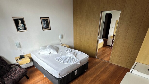 A modern bedroom with a large bed, a wooden floor, a bedside table with a lamp, and two framed pictures on the wall. - Hotel St. John (Blumenau)